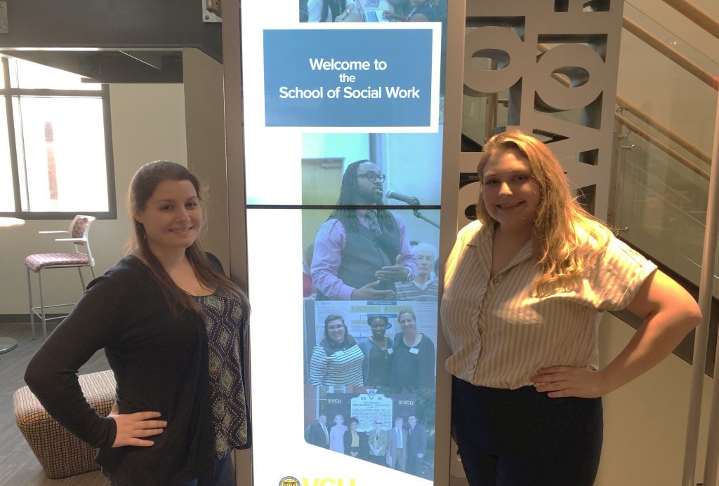 M-S-W students Elena Papathanassiou and Taylor Wilkerson stand smiling in front of signs for the VCU School of Social Work in the third floor lobby of the Academic Learning Commons building. 