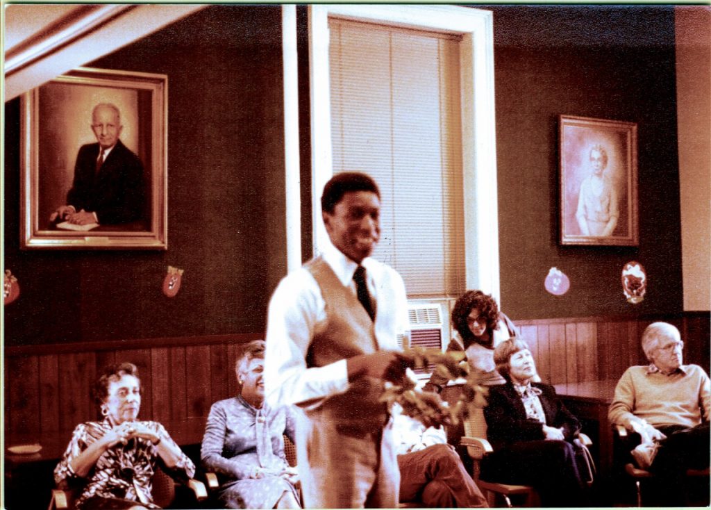 Bob Peay stands in front of seated colleagues from the School of Social Work. Two framed photos are on the wall behind him.