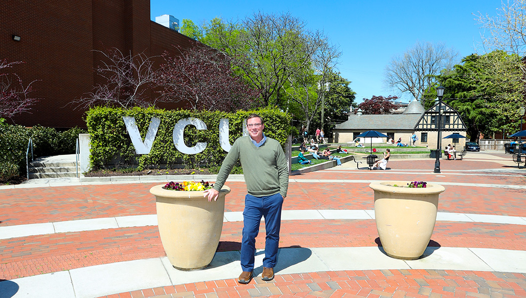 VCU School of Social Work administrator Gary Cuddeback leans against a large planter with colorful flowers, in front of a white VCU sign and a terraced green area where students are sitting.