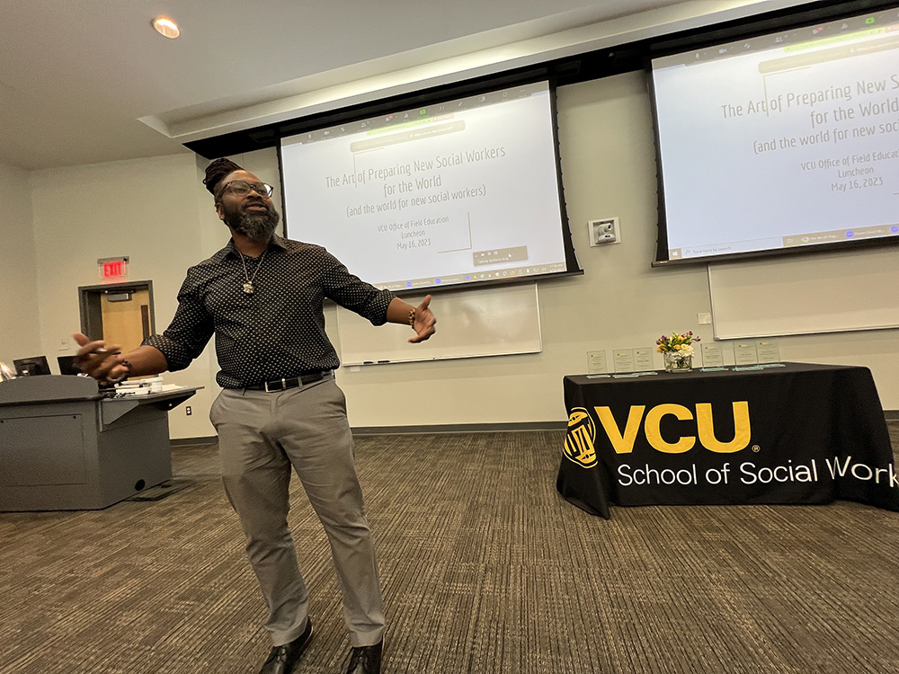 Alum Daryl Fraser raises his right hand to gesture as he talks inside a classroom. In the background, an attendee sits and listens; black, white and gold balloons are visible in the background.