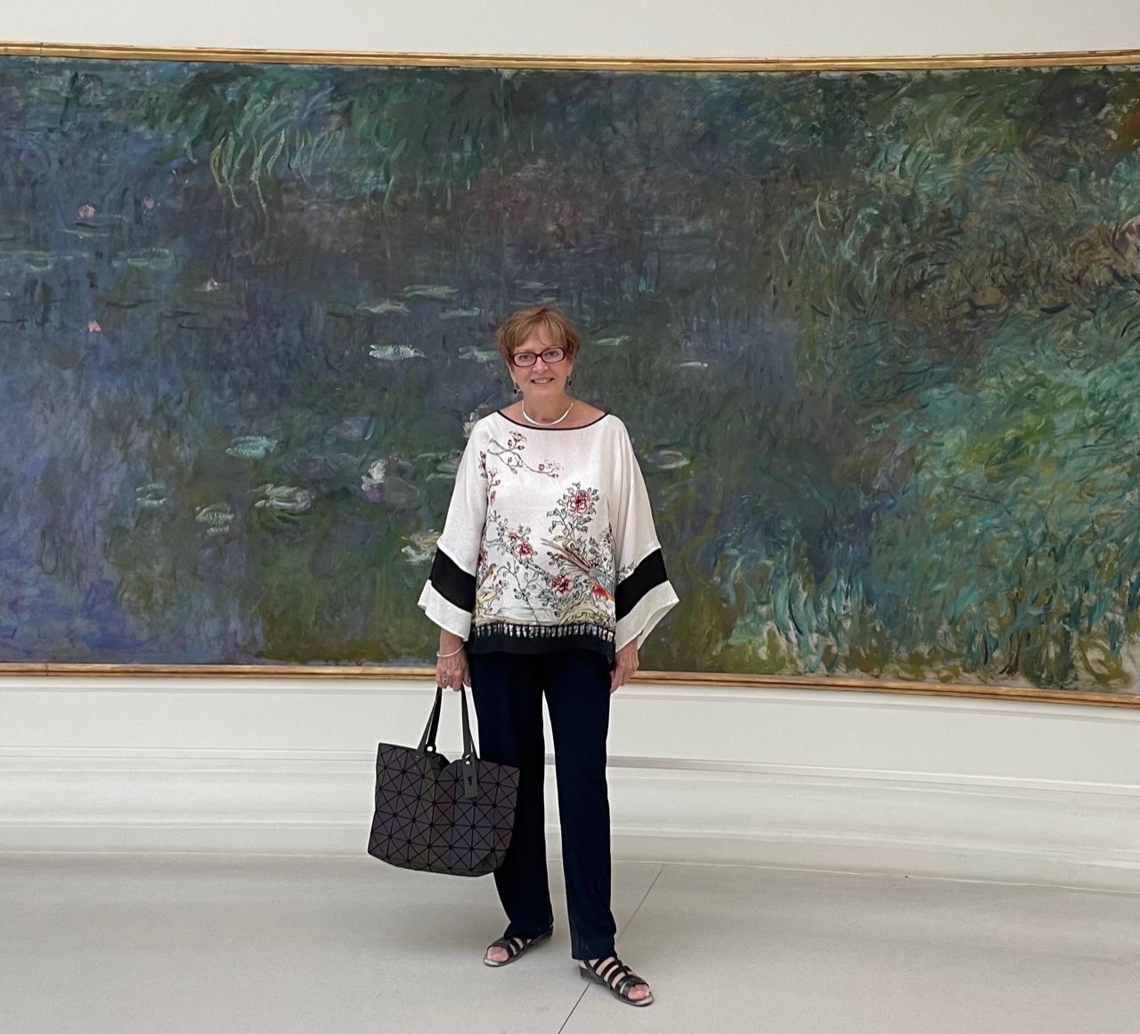Marjorie Stuckle stands in front of a large painting, Monet's "Water Lillies," with shades of purple, green, blue and brown. She is wearing a white print top, black pants, black sandals and holding a handbag.