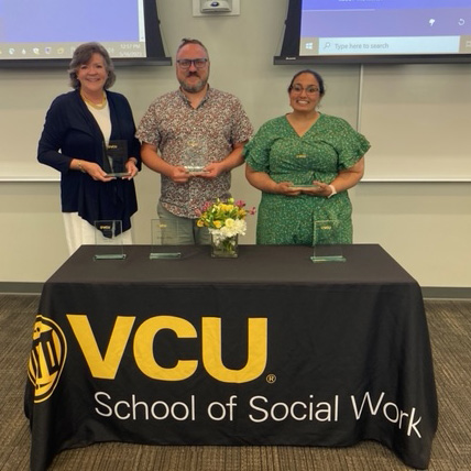 Three award recipients hold their awards, standing in front of a table with flowers and a black table cloth that reads "VCU School of Social Work."
