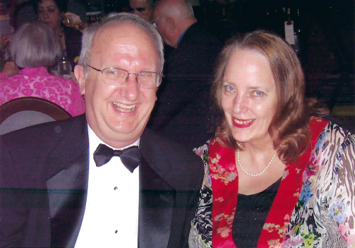 Seated next to other, Chuck Cartledge smiling and wearing a black tux and bowtie with a white shirt; and Mary Cartledge smiling and wearing a print jacket, black top and pearl necklace.
