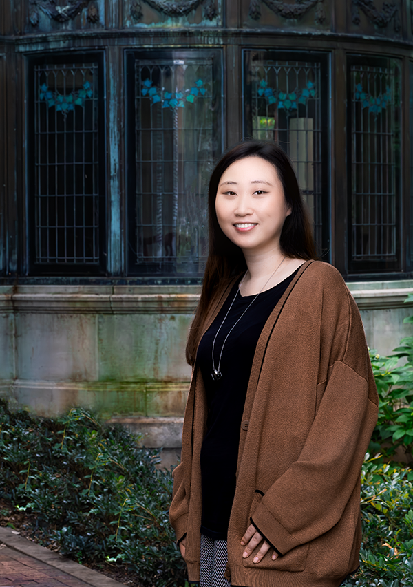 Doctoral student Katie Kim wears a black top, gray pants and a brown sweater. She stands outside a stone and glass building exterior flanked by greenery.