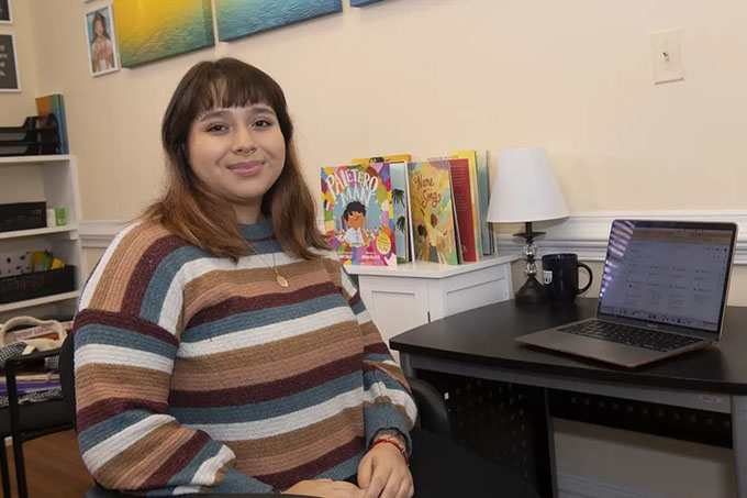 Paloma Rodriguez sits in an office in front of a desk with a laptop computer and children's books in the background. 