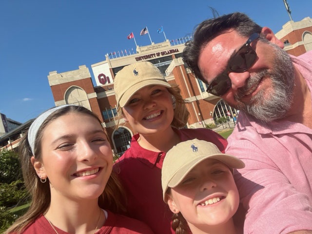 Family of four poses, smiling, in front of a football stadium. 