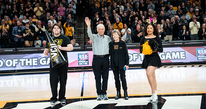 Flanked by a VCU cheerleader with a megaphone and one with black and gold pompoms, Dr. Frank Baskind stands at center court of the Siegel Center basketball court and waves while putting his arm around his wife, Patricia. 