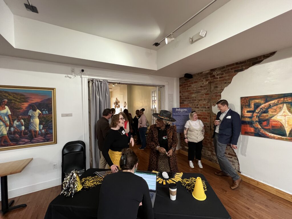 Alumni, students and staff stand in front of a registration table draped in a black cloth. Colorful artwork hangs on the wall, part of which is exposed brick.