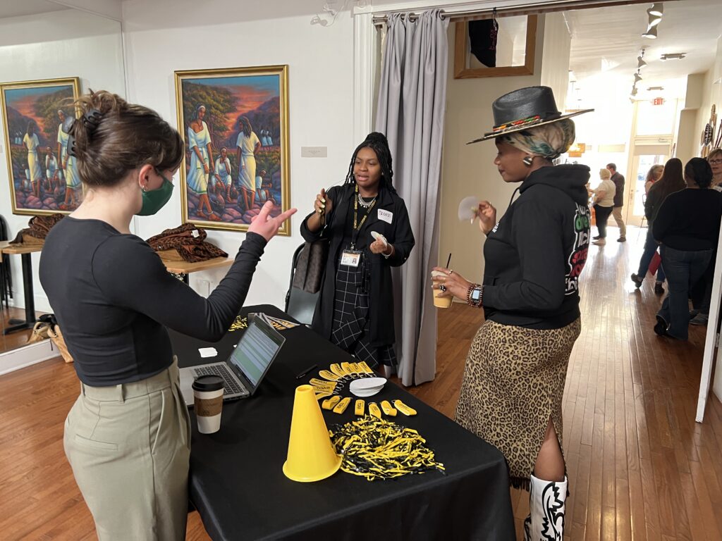 Three people stand around a registration table draped in a black cloth on top of which are a gold megaphone and black and gold pompoms. 