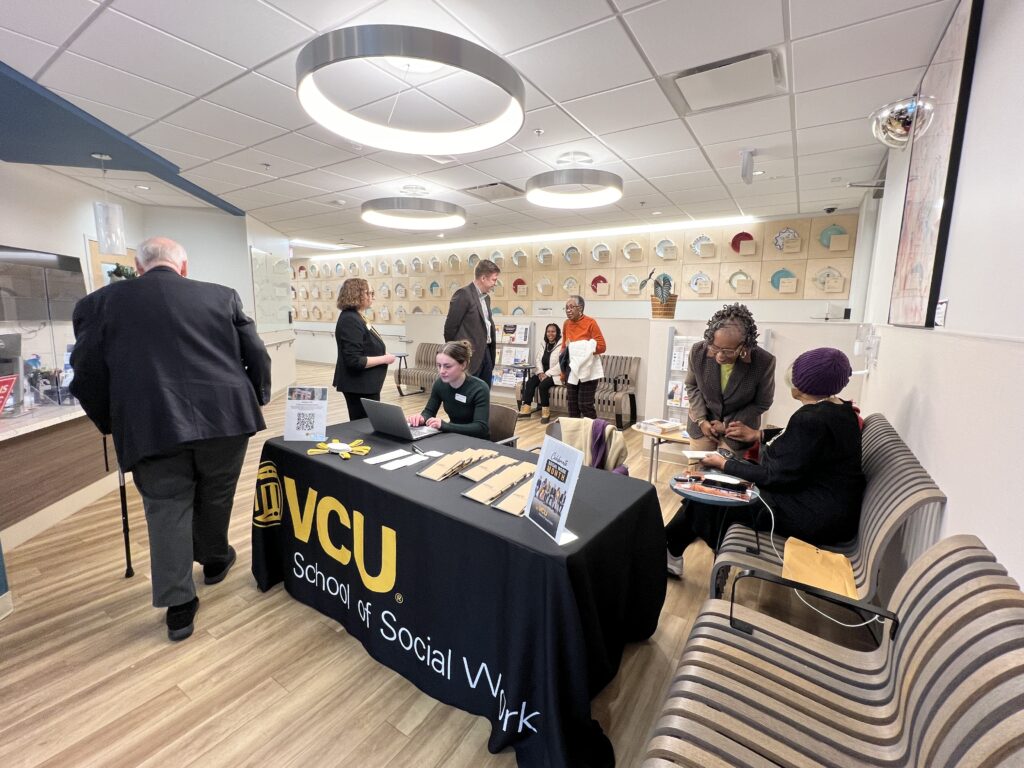 People stand, walk, sit and chat around a registration table draped with a black cloth that reads VCU School of Social Work