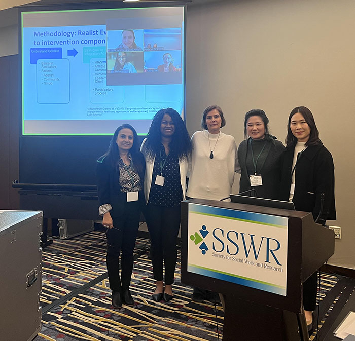 Five presenters stand behind in front of a presentation screen that reads in part Methodology: Realist ... to intervention ...; and in front of a podium that reads SSWR - Society for Social Work and Research. 