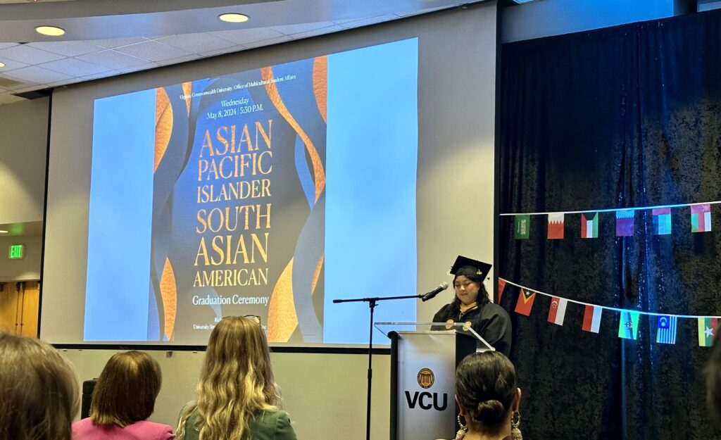 A student in black graduation cap and gown stands at a podium and speaks. A digital screen reads: Asian Pacific Islander South Asian American. To the side are small flags of various international countries.