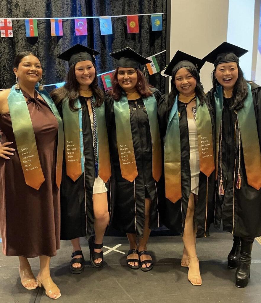 Five students, four in black graduation caps and gowns, stand with stoles reading: Asian Pacific Islander South Asian American. In the background are small flags of various international countries.