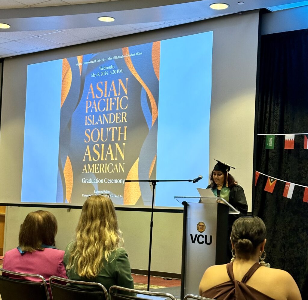 A student in black graduation cap and gown stands at a podium and speaks. A digital screen reads: Asian Pacific Islander South Asian American. To the side are small flags of various international countries.