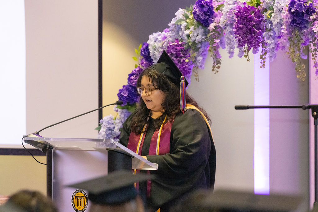 A student in black graduation cap and gown stands at a podium and reads. Behind are an array of purple and lavender flowers.