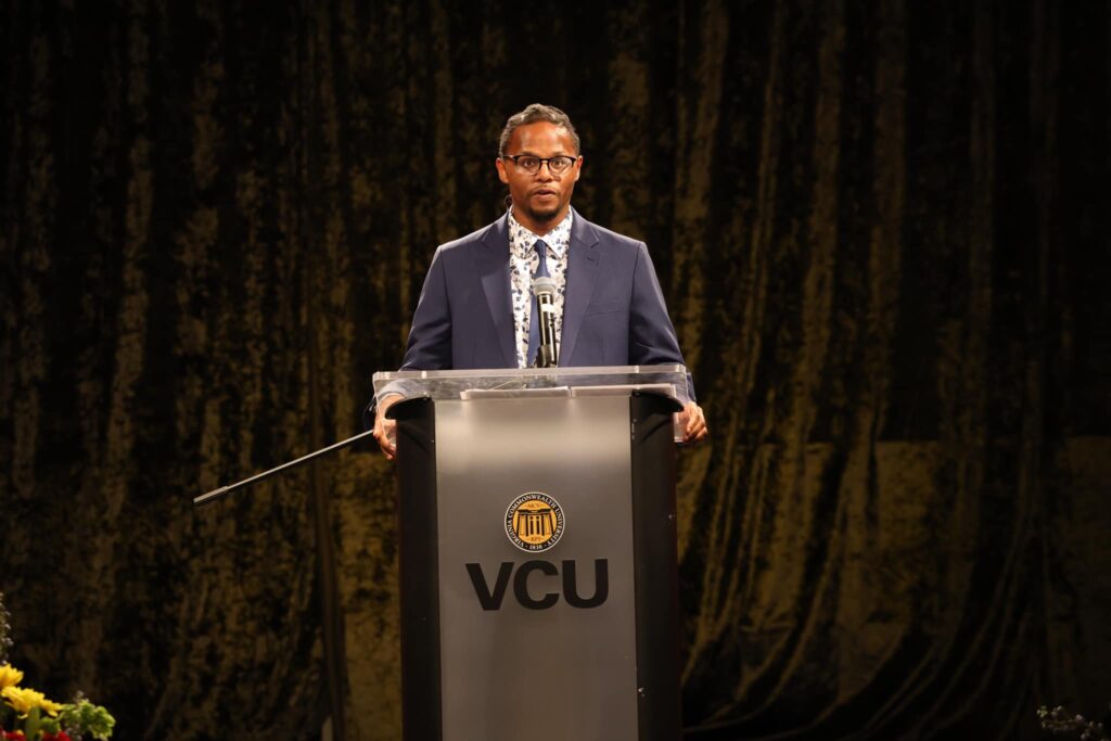 A man in a dark suit and tie stands and speaks behind a podium with a sign reading: VCU.