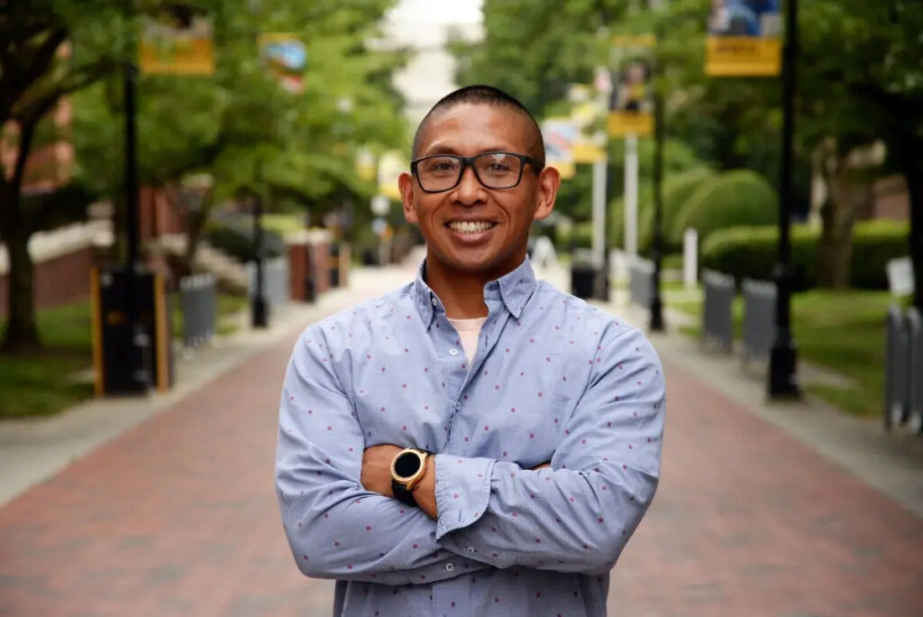 Portrait of Samuel Ochinang in blue shirt with arms crossed, standing on a brick pathway. 