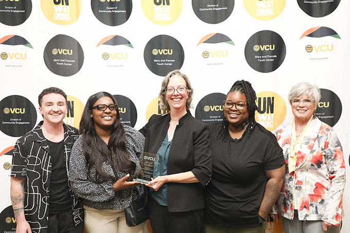 A group of five people stand with two in the center holding an award. In the background are graphics that say VCU Division of Community Engagement and "Unwavering."