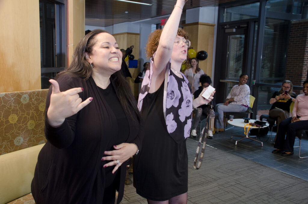 A staff and faculty member gesture and smile as they support students performing karaoke.