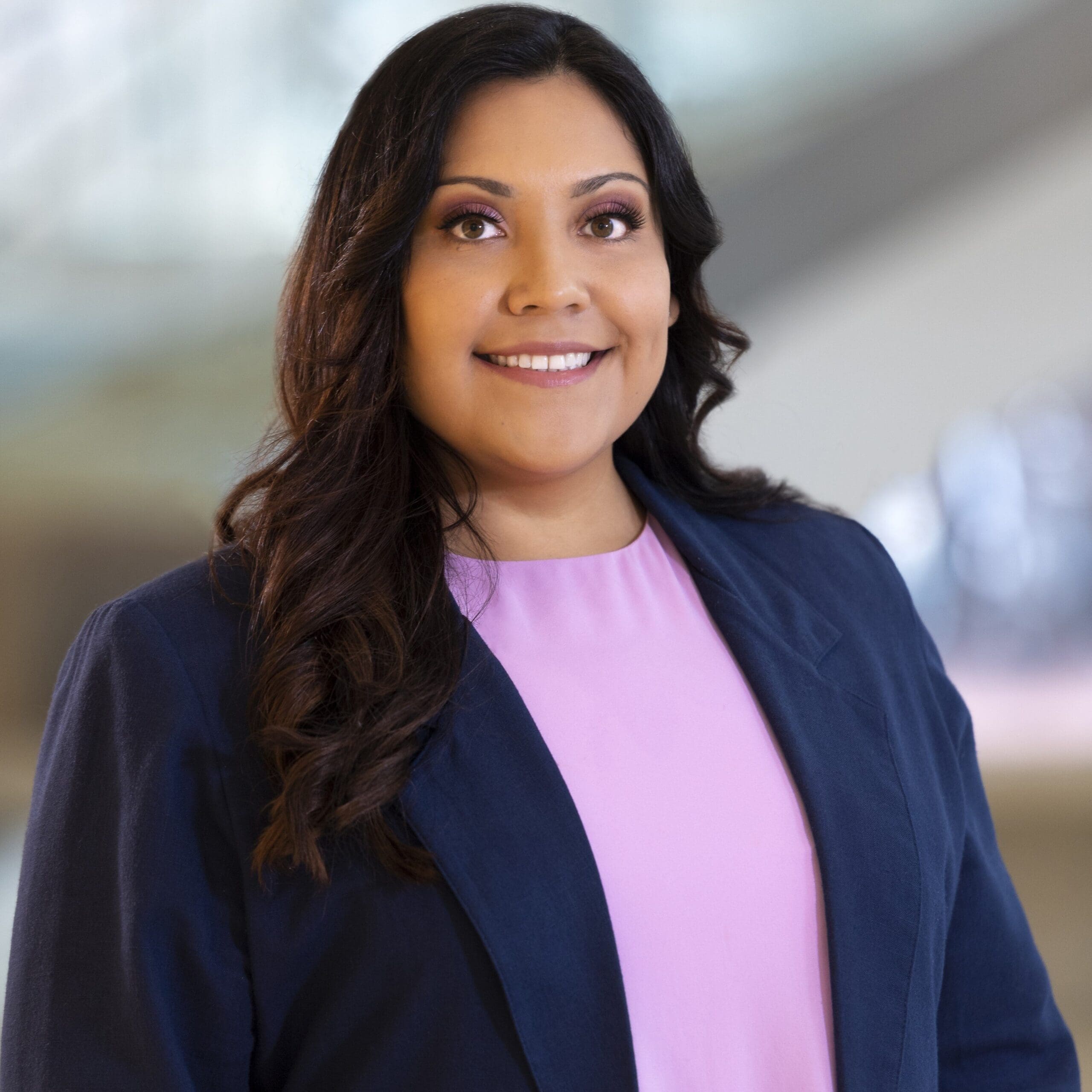headshot of Dina Garcia, wearing a pink shirt with a dark blue blazer. she is smiling her hair is brown and wavy.