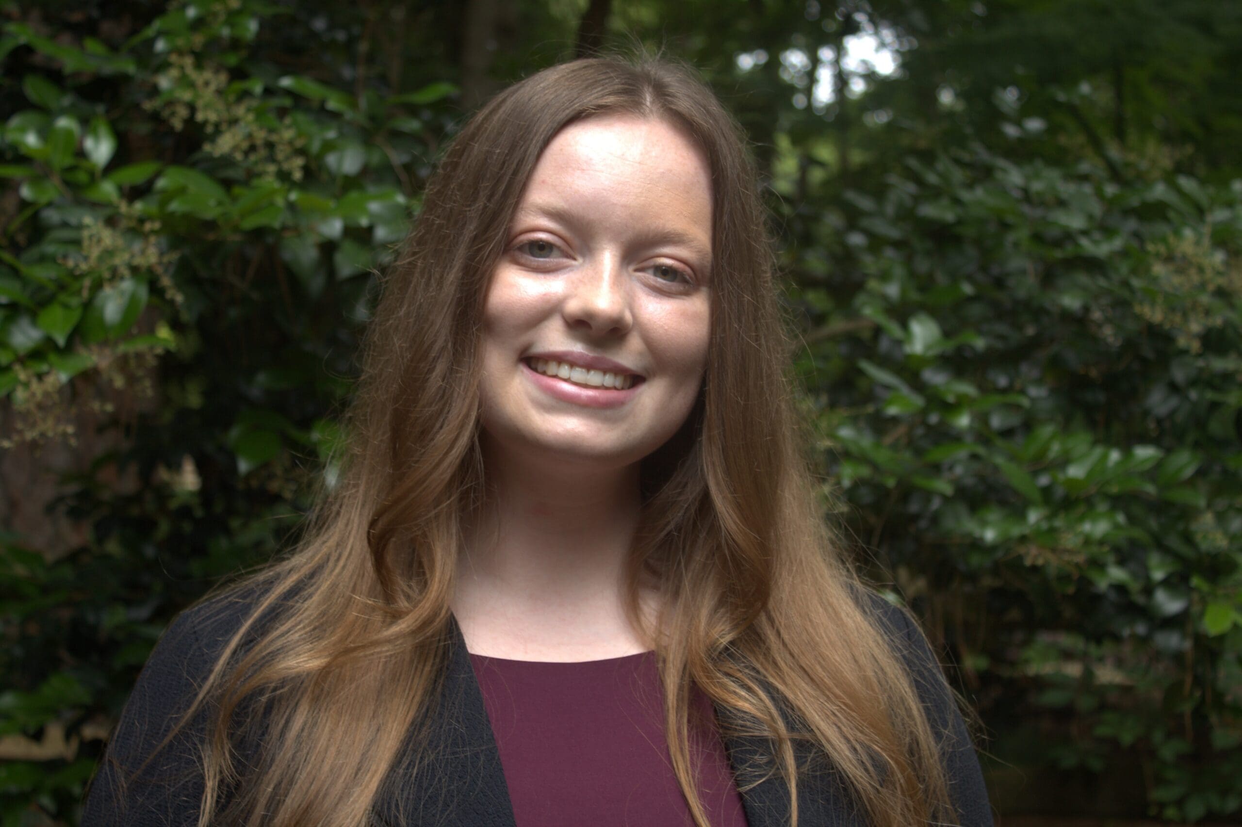 a young women with light brown hair is smiling. she is outside in front of greenery, wearing a maroon shirt and black sweater