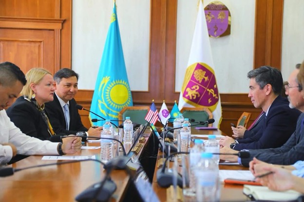 Five people in business attire sit at a long table with water bottles and microphones between them. Two flags, including the flag of Kazakhstan are in the background.