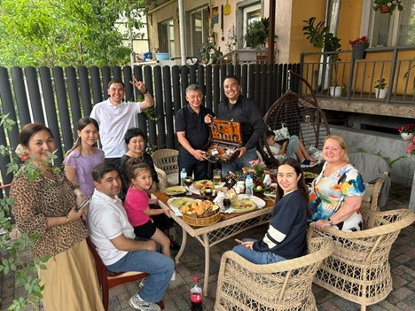 A group of nine adults and two children sit at a table outside enjoying food and smiling.