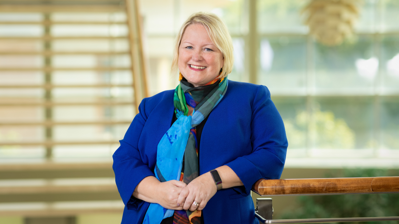 a middle-aged woman with blond hair is smiling with her hands folded, wearing a blue blazer and a bright blue and gree scarf.