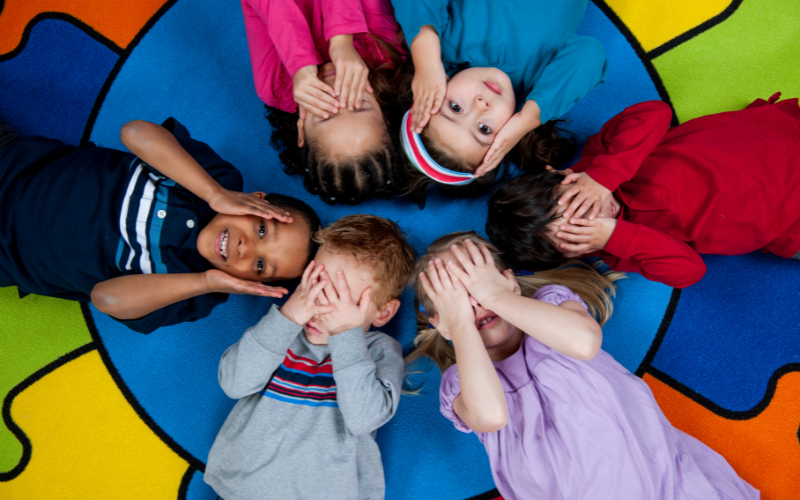 Group of children laying in a circle in thhe rug covering their eyes and ears