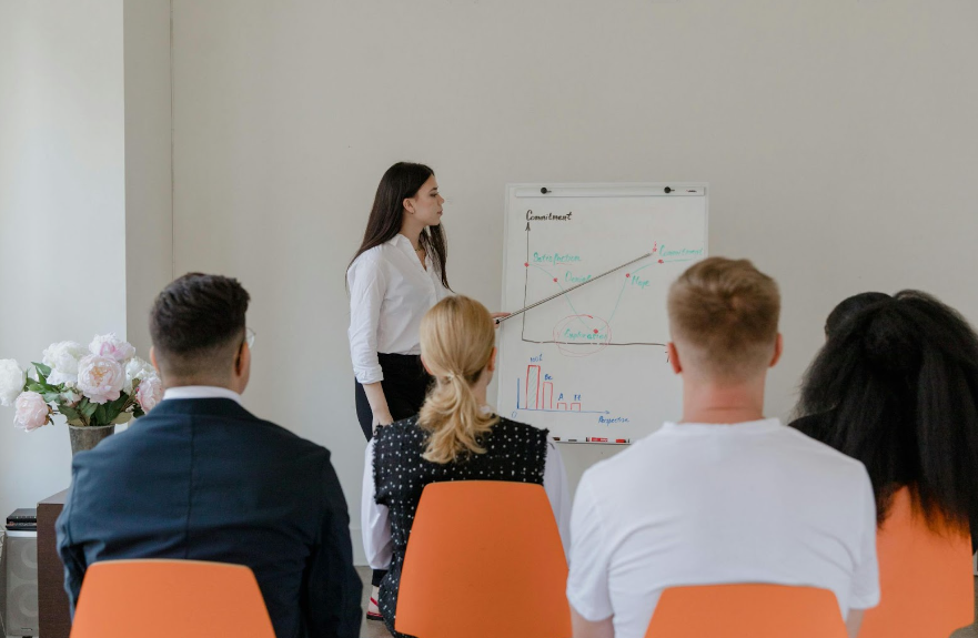 Image of a woman standing at a white board with a pointer. There is a group of adults watching her present.