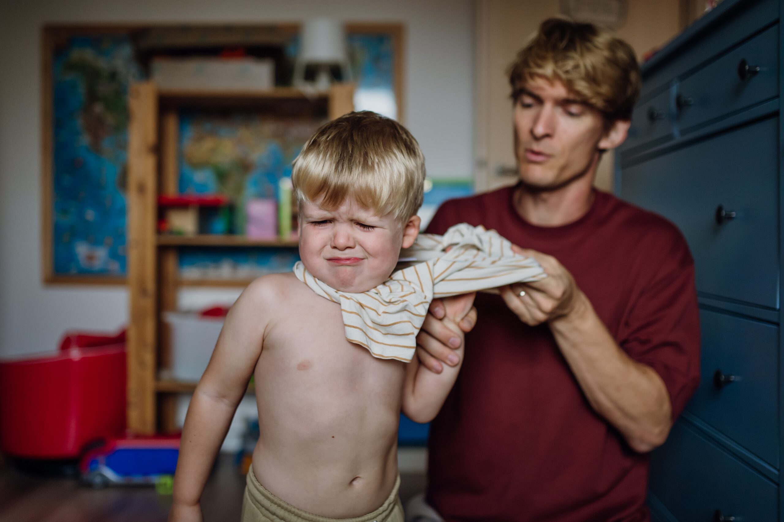 Father putting shirt on little son, changing from pajamas in the morning. Boy is sad, crying.