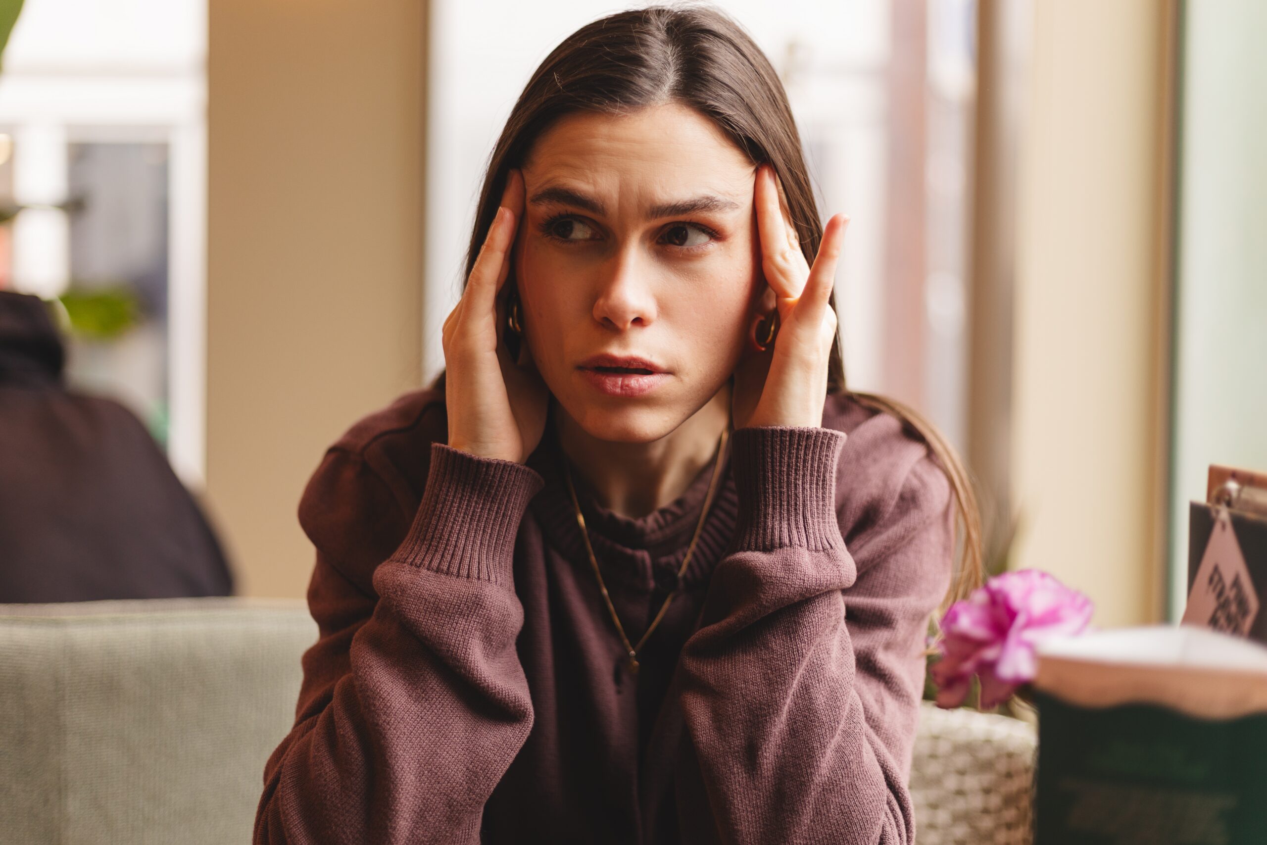 Stressed young woman suffering from a painful headache sitting in a cafe with her hands on temples and looking away with a worried facial expression due to a sudden migraine or tension
