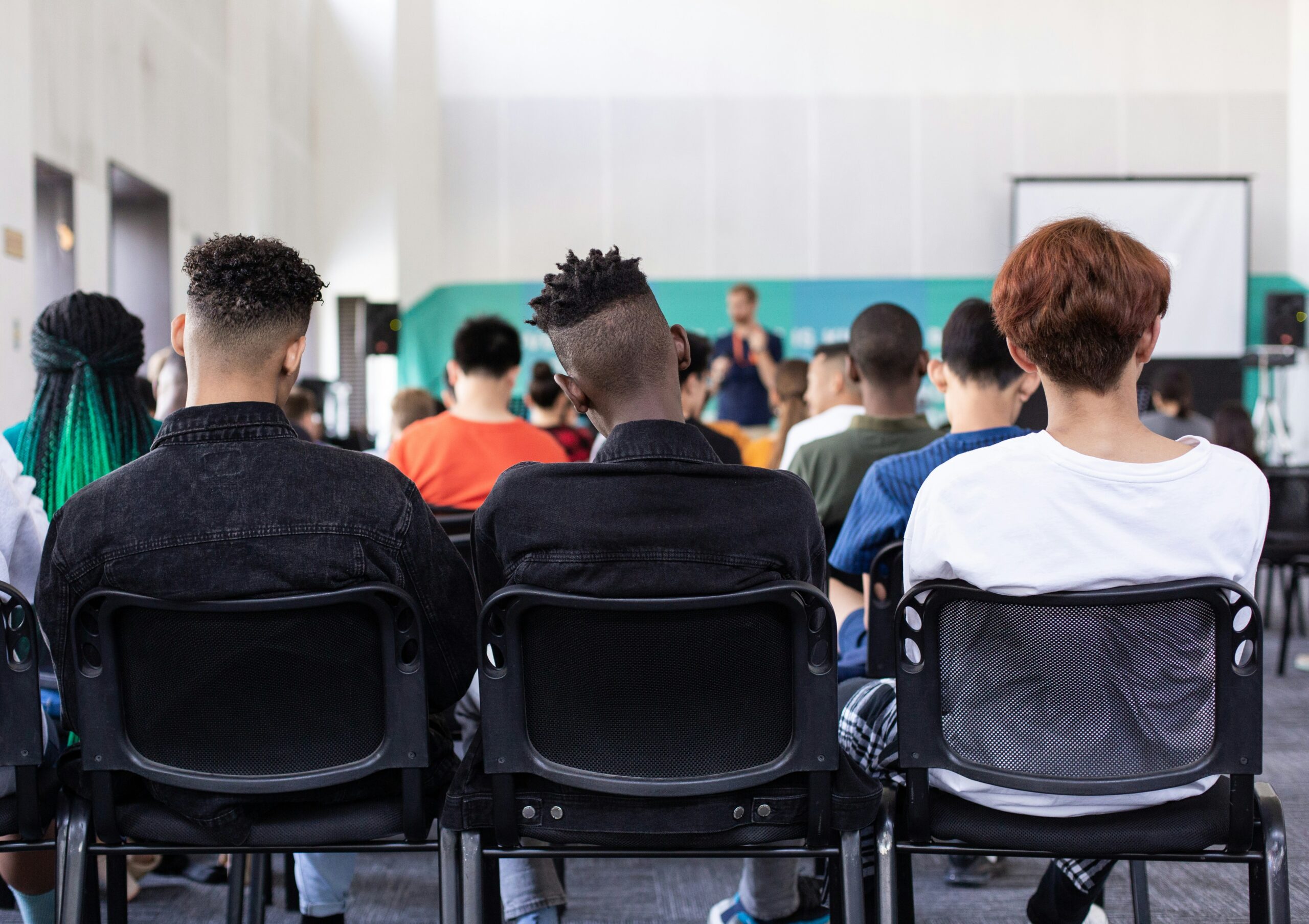 Three people sit in chairs in the back of a full lecture hall. Only the backs of their heads are visible.