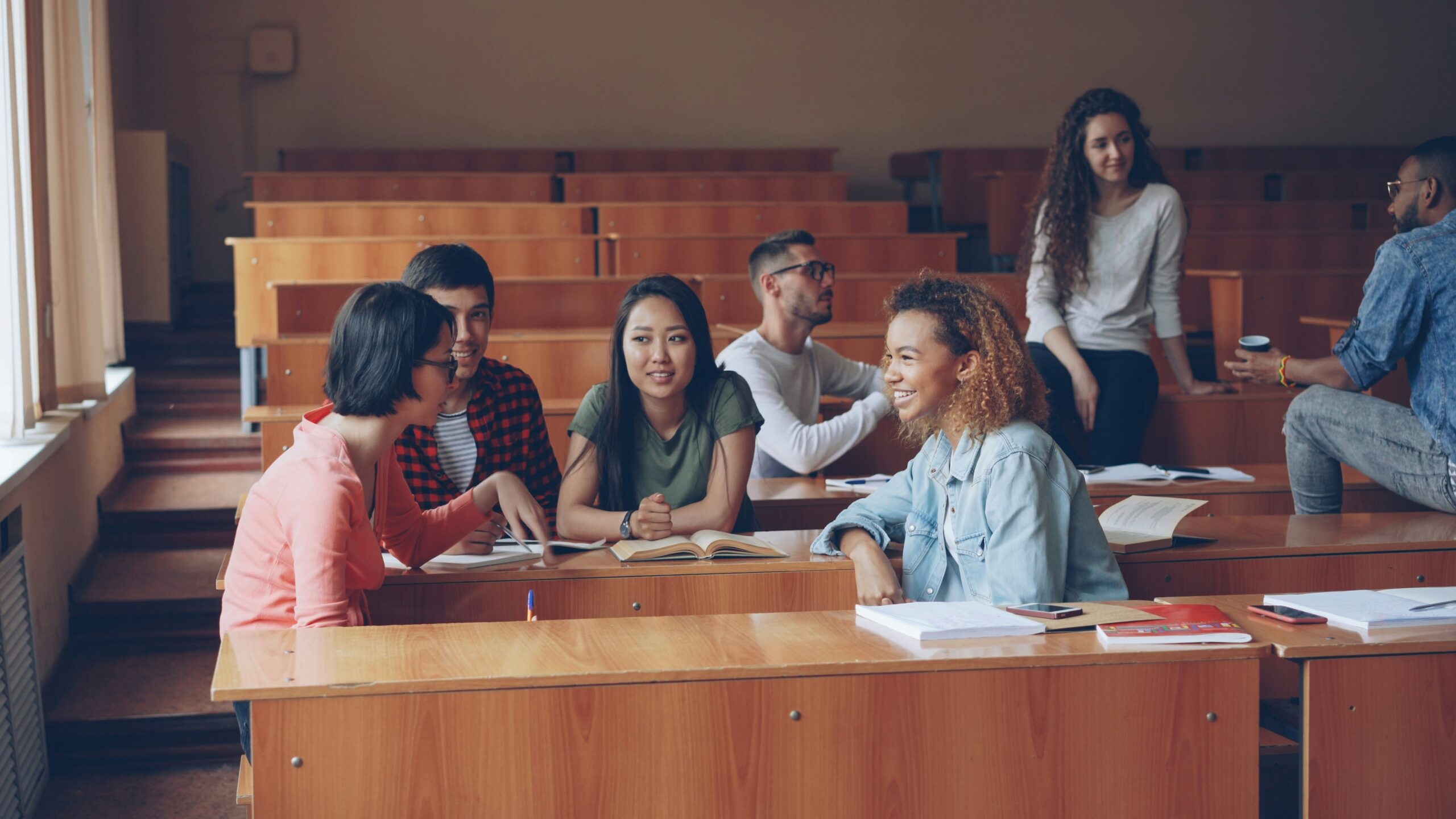 Multiracial group of university students are relaxing and chatting during break enjoying free time and communication. Wooden tables, attractive people and noteboks are visible.